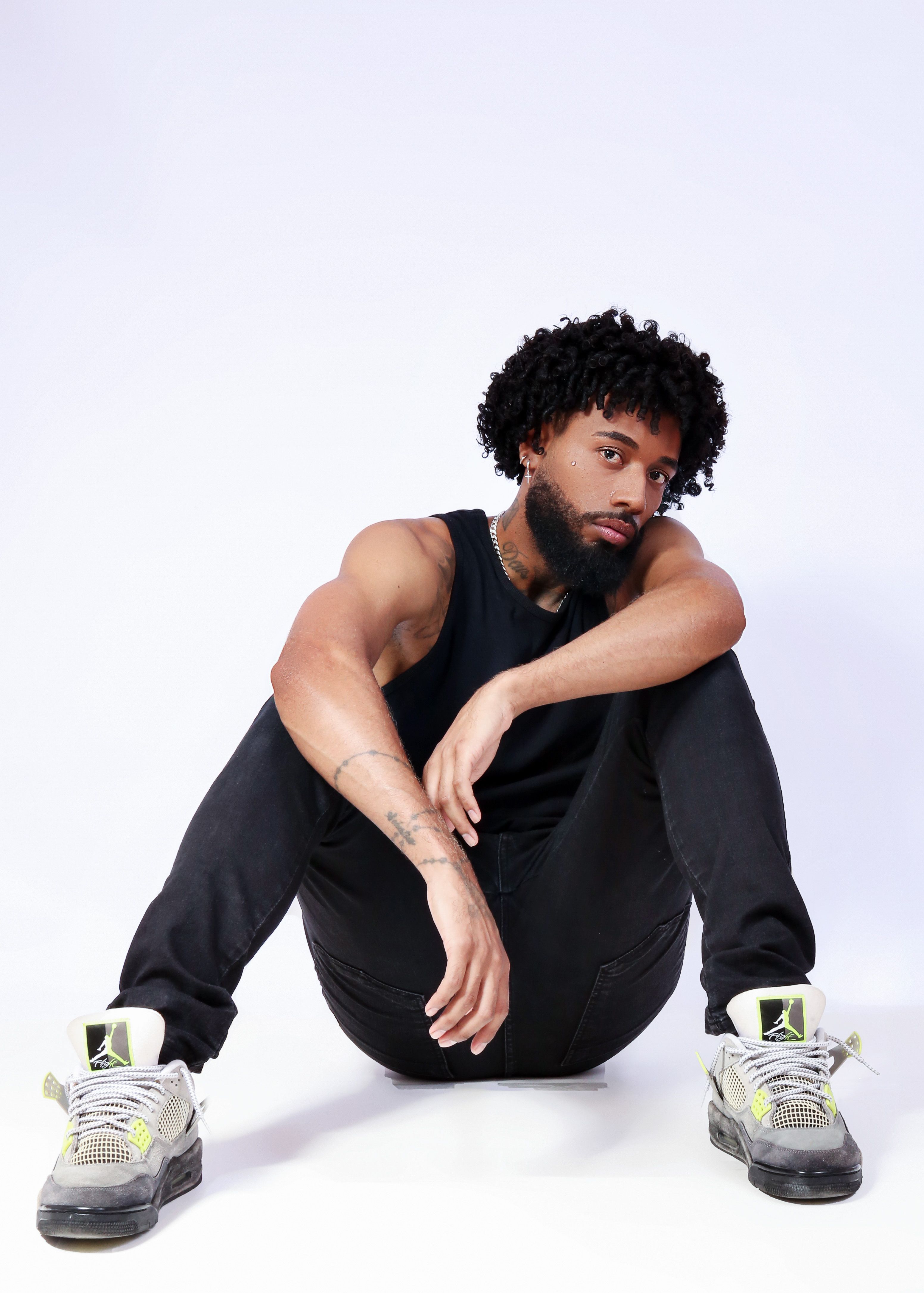 A fashionable man with curly hair and a beard poses in a black outfit against a white background, embodying modern style.