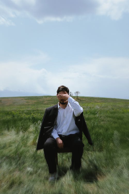 Elegant Man Crouching in Field under Blue Sky Covering Face with Hand