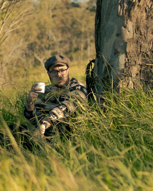 Free A man wearing a sweater relaxes against a tree, enjoying coffee in a sunny meadow. Stock Photo