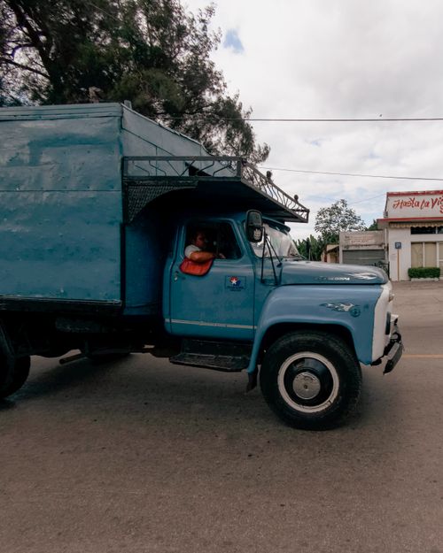 Free Blue vintage truck driving down a street under an overcast sky in an urban setting. Stock Photo