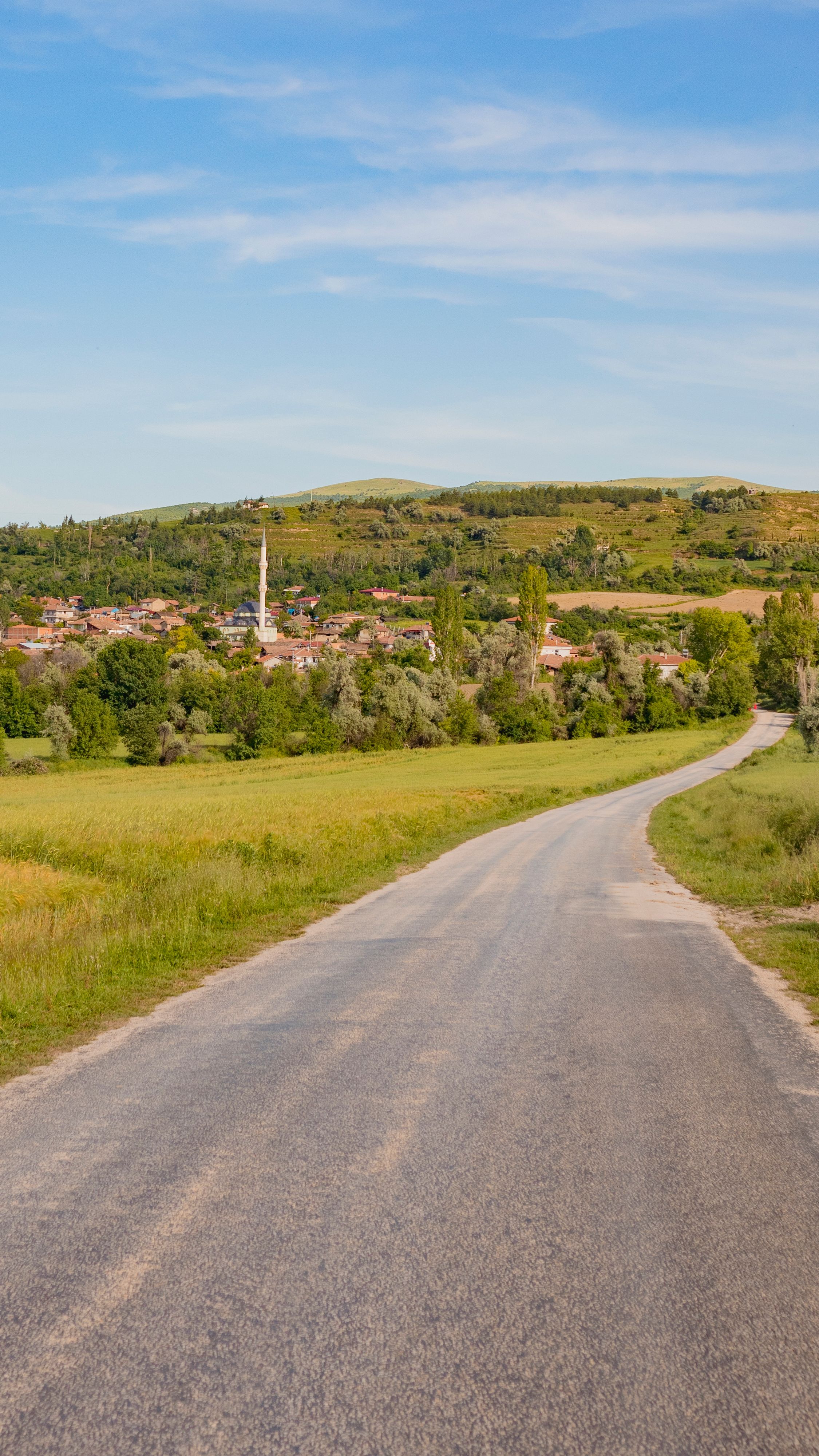 A sunlit country road winding through rolling hills towards a distant village, surrounded by greenery.