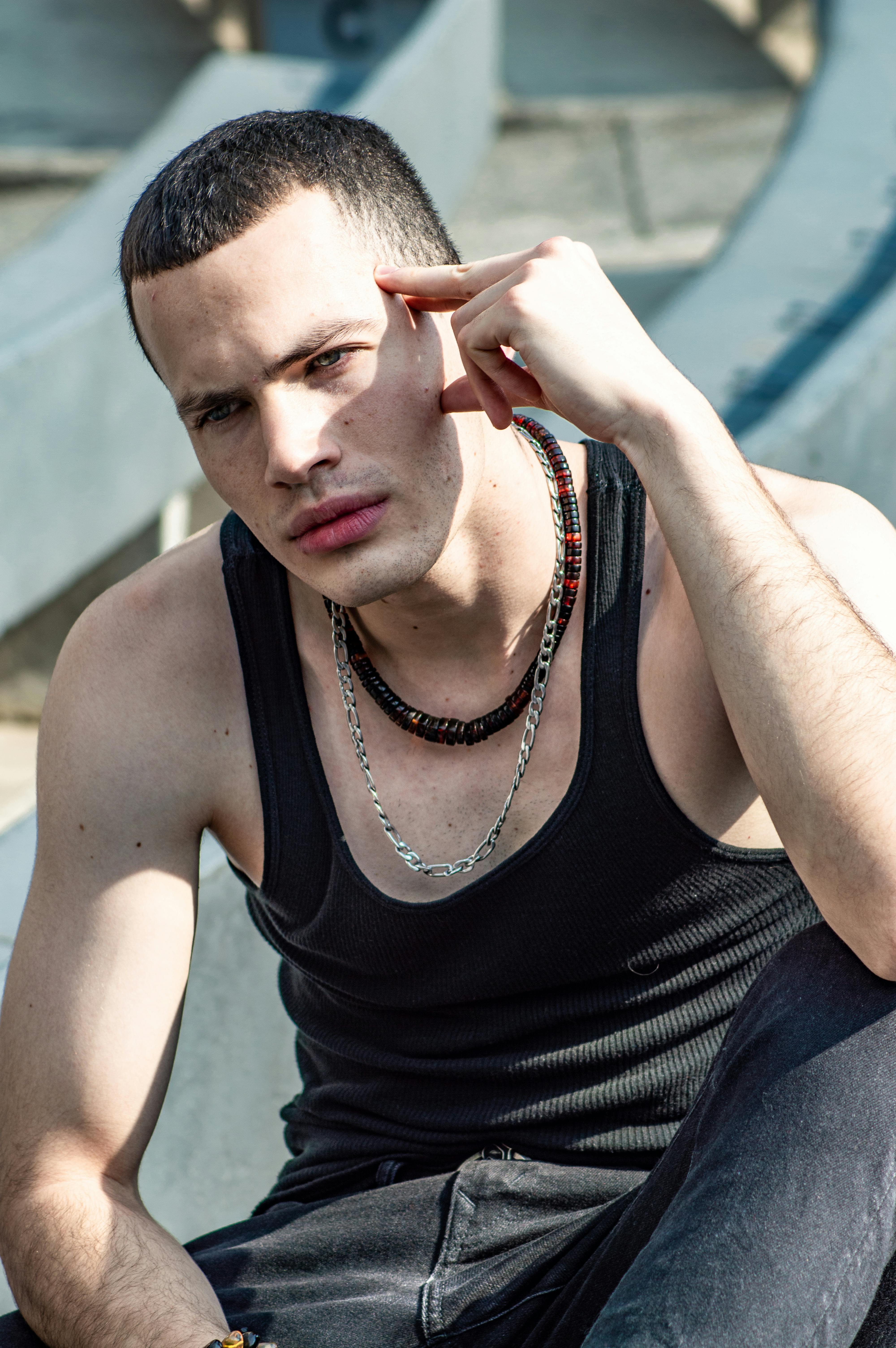 Stylish male model in black tank top sitting outdoors in sunlight.