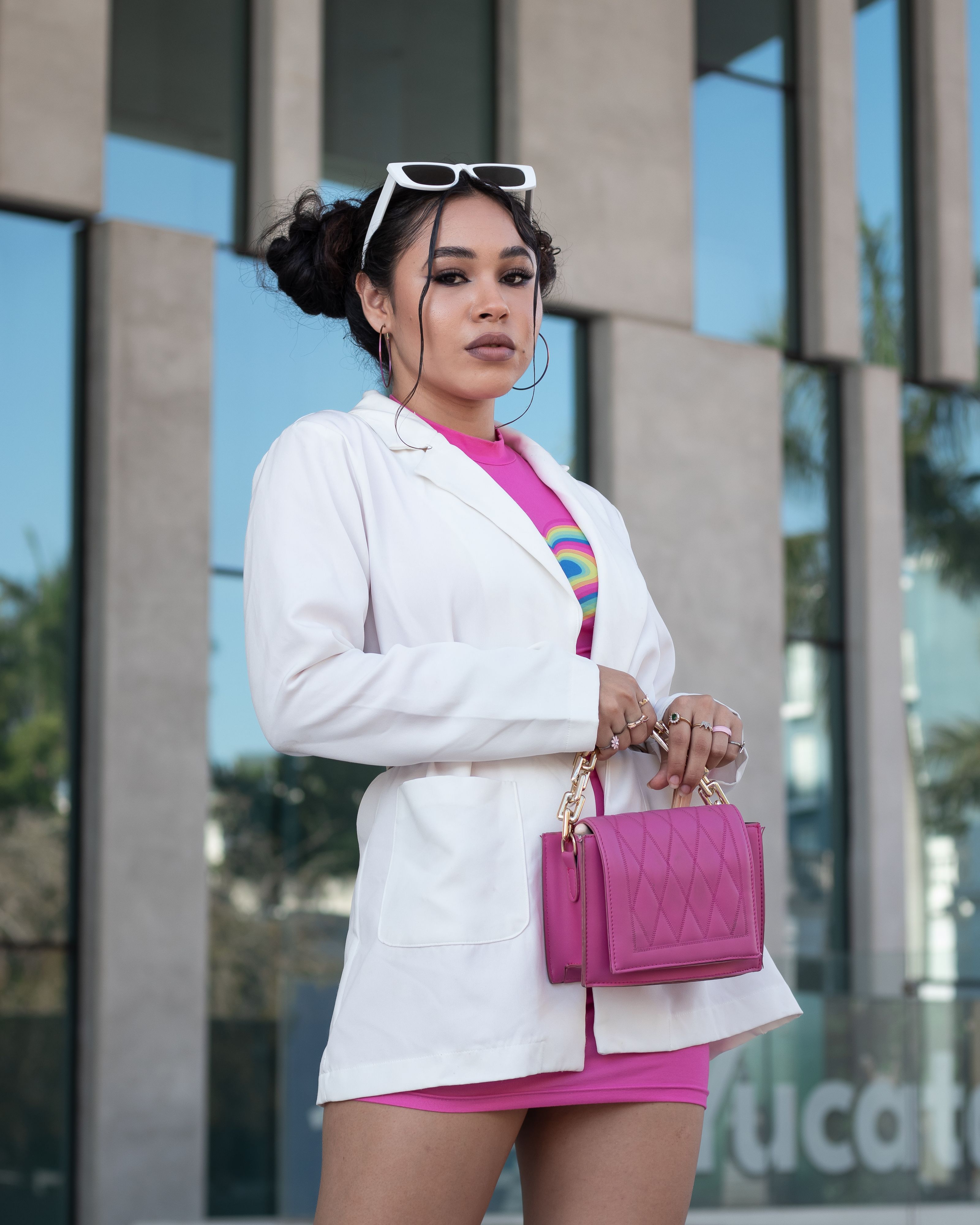 Mulher estilosa com uma bolsa rosa posa confiantemente ao ar livre vestindo um blazer branco e roupas coloridas.