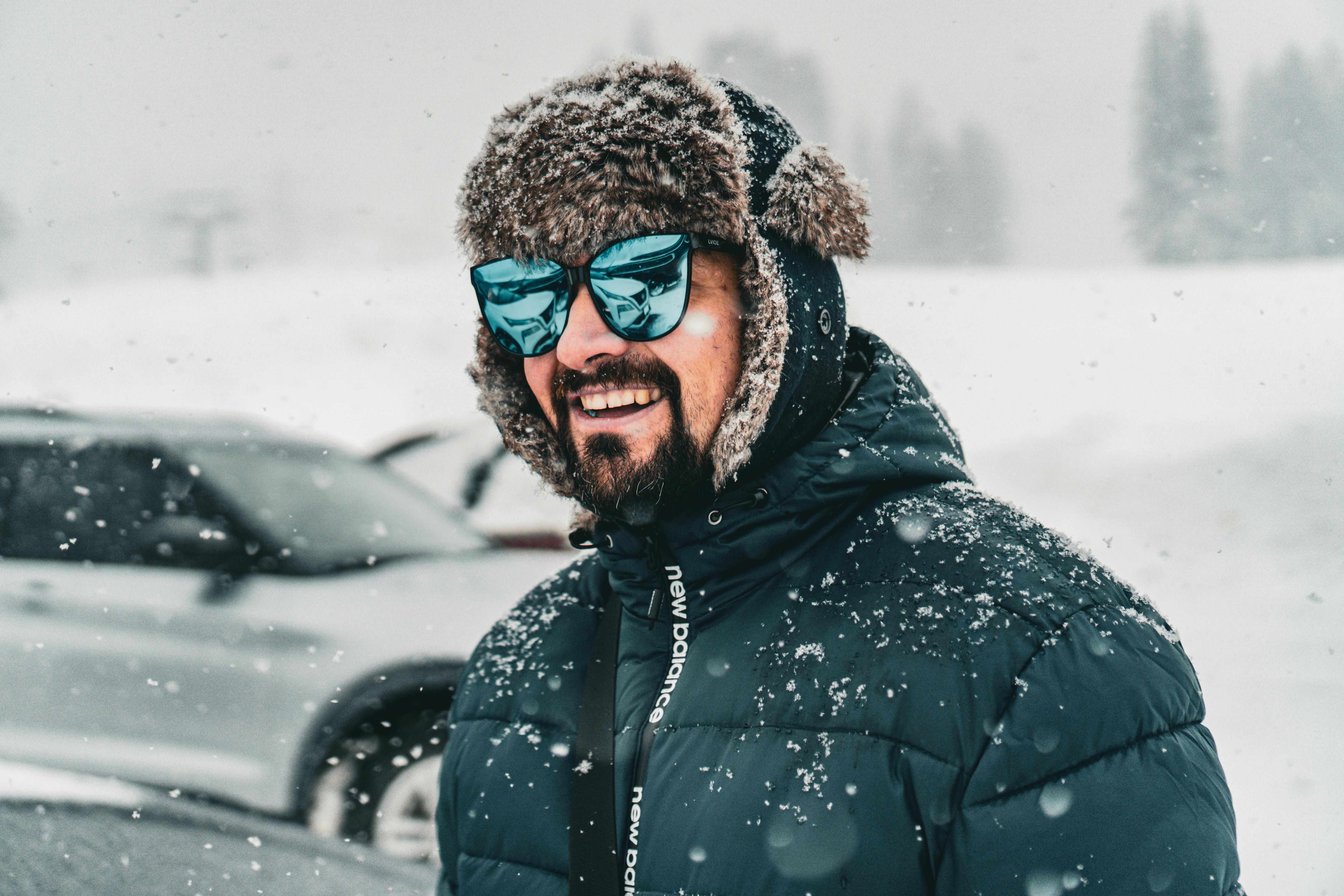 Man in winter attire smiling in snowfall, wearing an ushanka and reflective sunglasses.