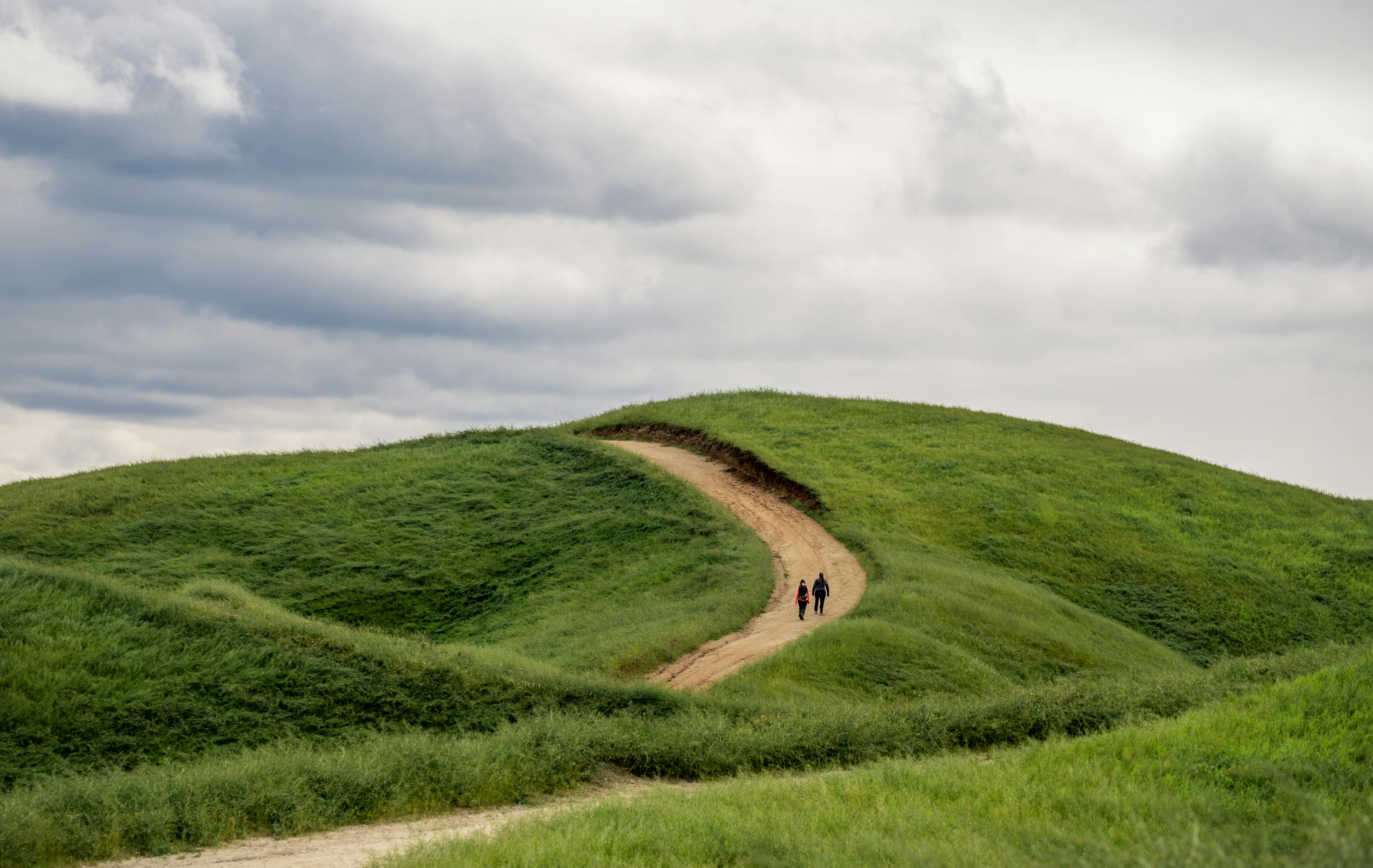 Two hikers traverse a winding trail on lush green hills under a cloudy sky in Loma Linda, California.