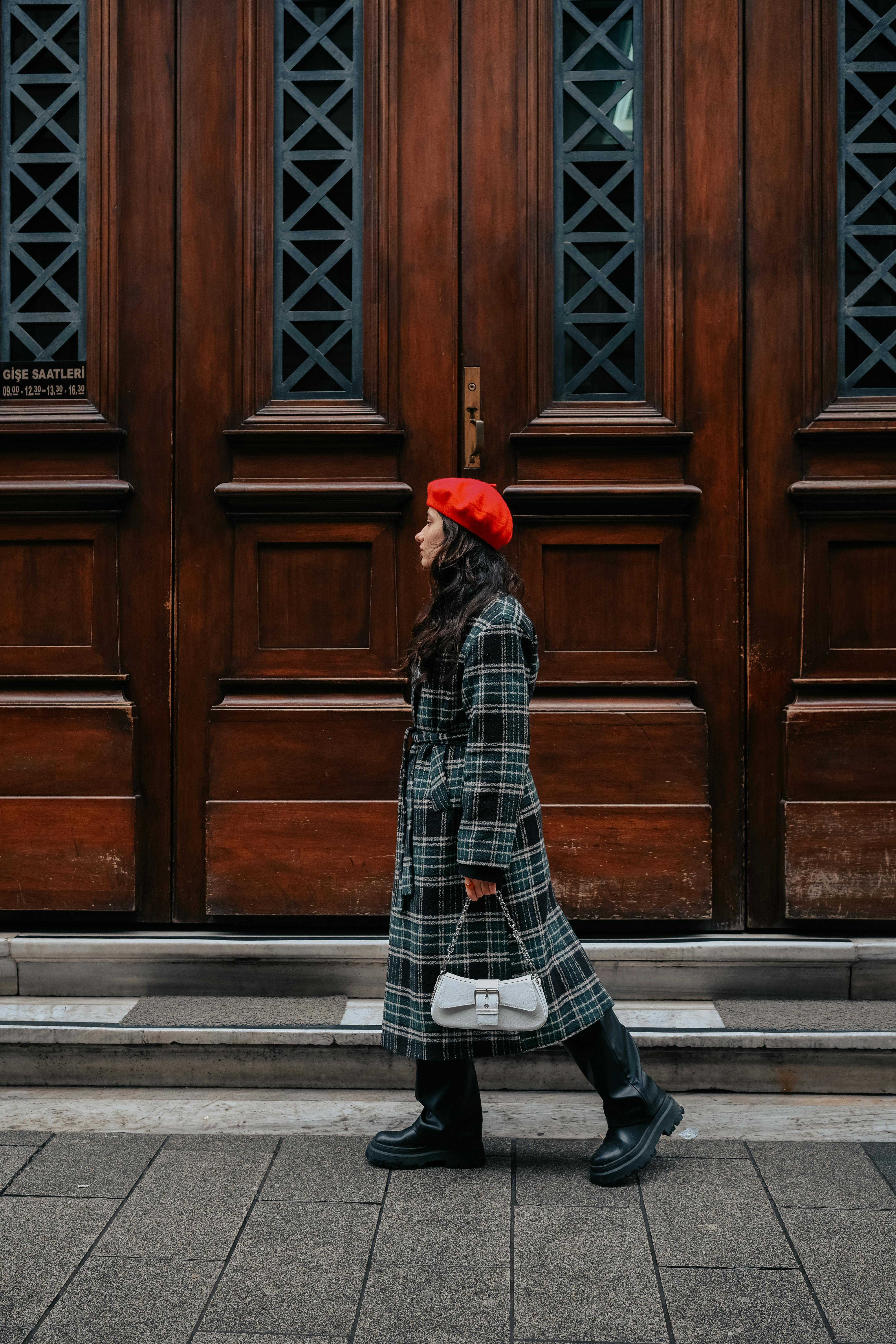Stylish woman in a checkered coat and red beret walking beside a wooden building facade.