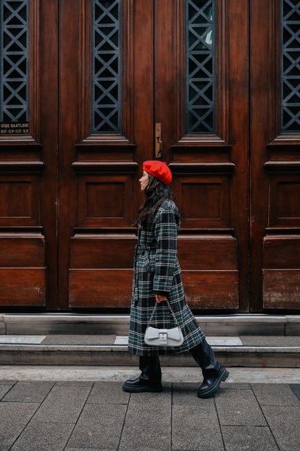 Stylish woman in a checkered coat and red beret walking beside a wooden building facade.