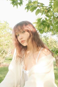A serene portrait of a woman with brown hair, wearing a white outfit under lush green trees.