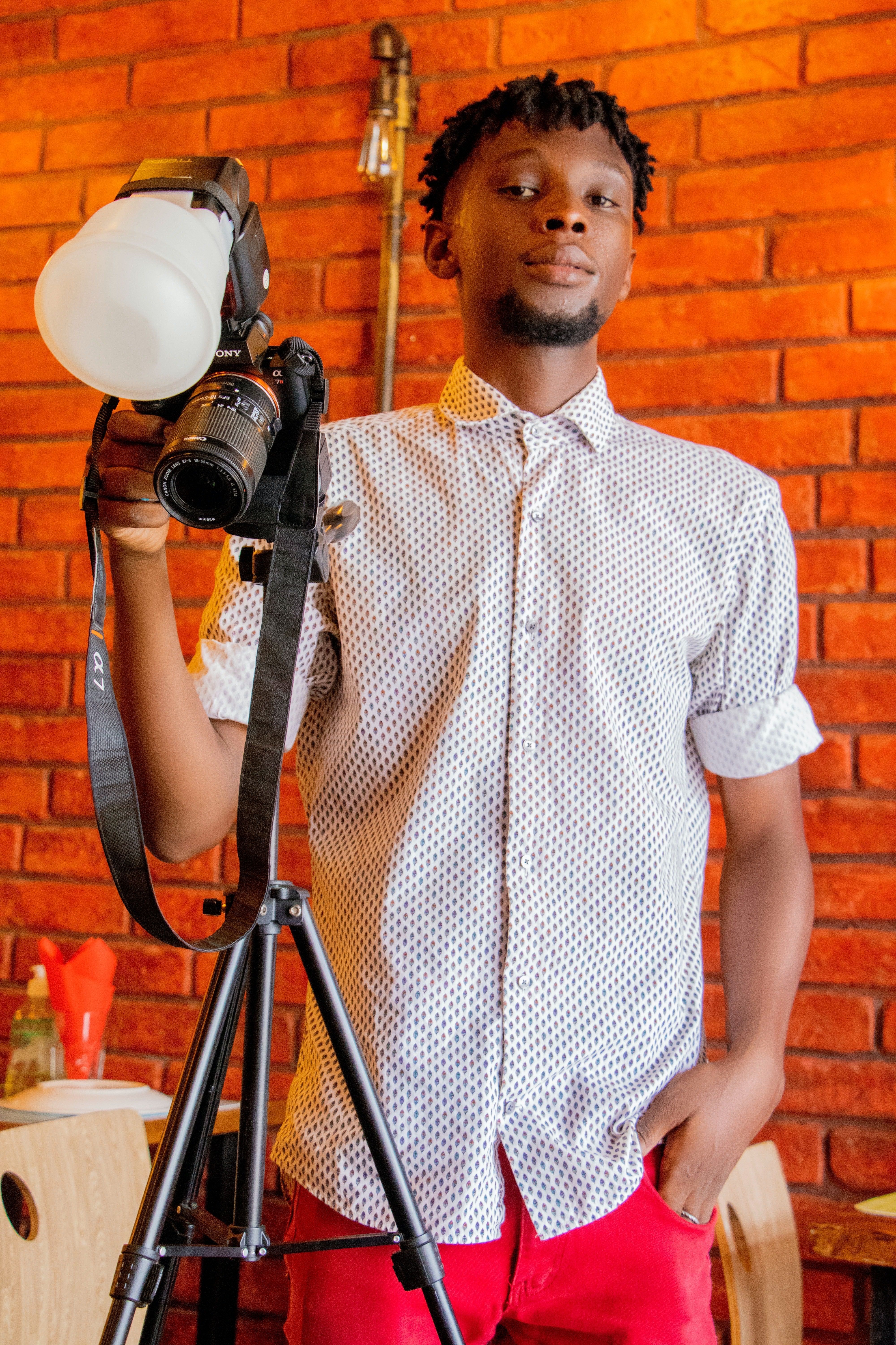 Young male photographer posing with camera and tripod against brick wall.