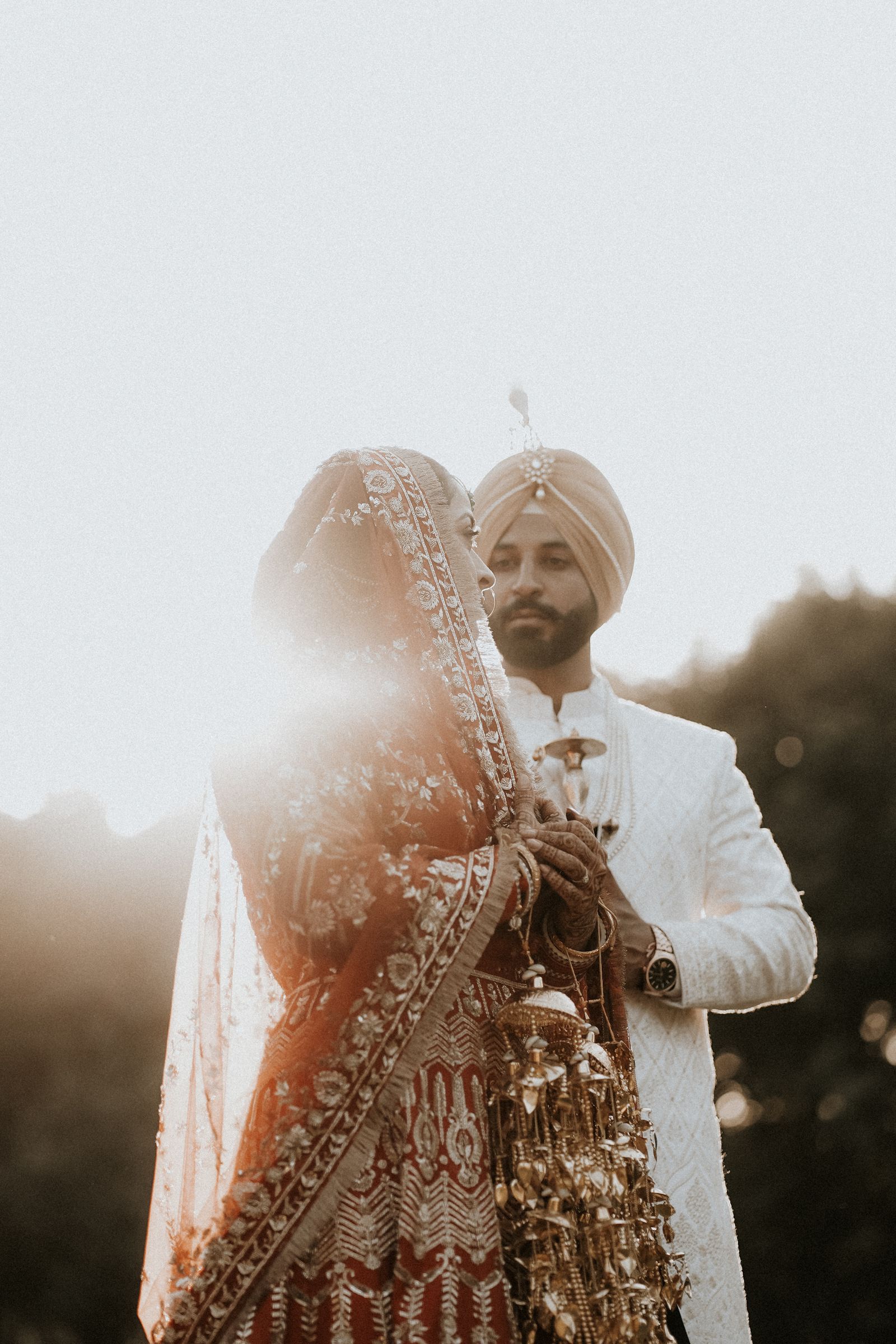 A couple standing into a low backlit sun, her red dupatta drawn over her head, gold kaleere hanging from her wrist, the sky above them blown to white.