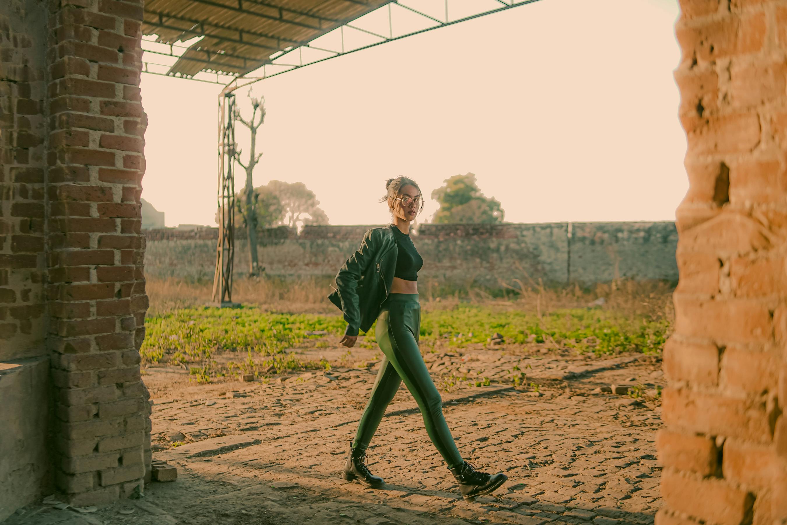 A woman in black attire confidently walks through an abandoned urban setting, lit by warm sunset tones.