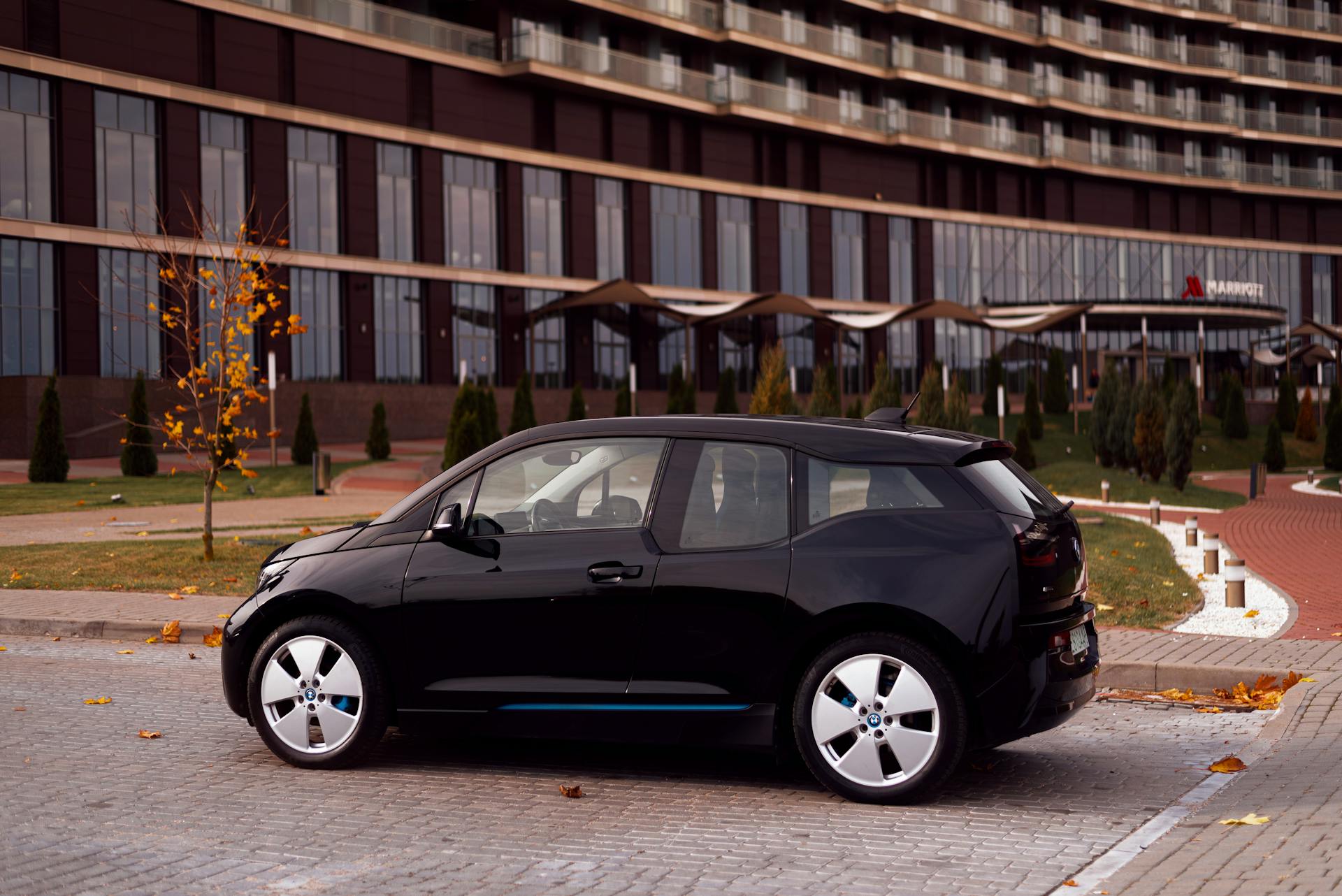 Black BMW i3 parked on city street with modern building backdrop, autumn.