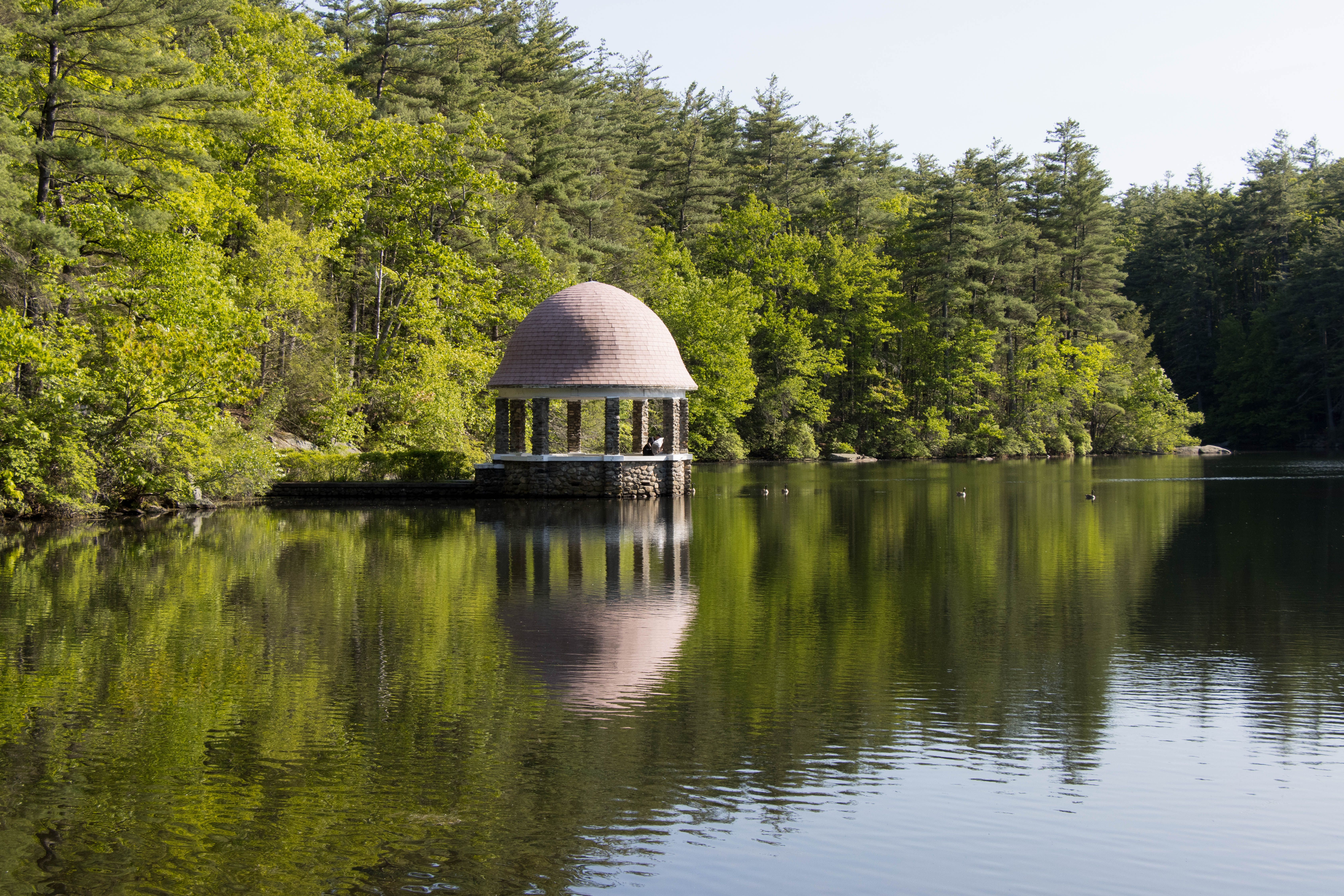 Tranquil scene of a gazebo reflecting in a pond surrounded by lush trees in Fitchburg, MA.