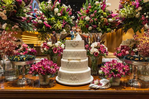 Free Three-tier white wedding cake surrounded by vibrant flower arrangements indoors. Stock Photo