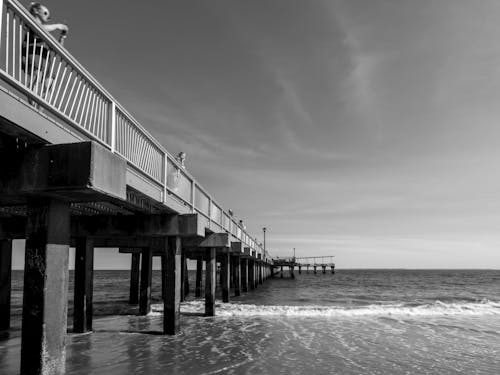 Black and White Photo of a Pier and Waves Washing Up the Shore 