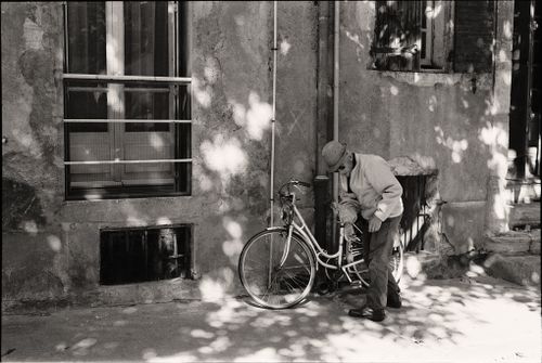 Free Black and white photo of an elderly man standing by a bicycle against a building wall. Stock Photo