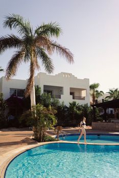 Young woman in bikini walking by a tropical resort pool, surrounded by palm trees.