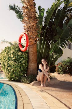 Woman sitting by a tropical poolside with palm trees, enjoying a relaxing vacation moment.