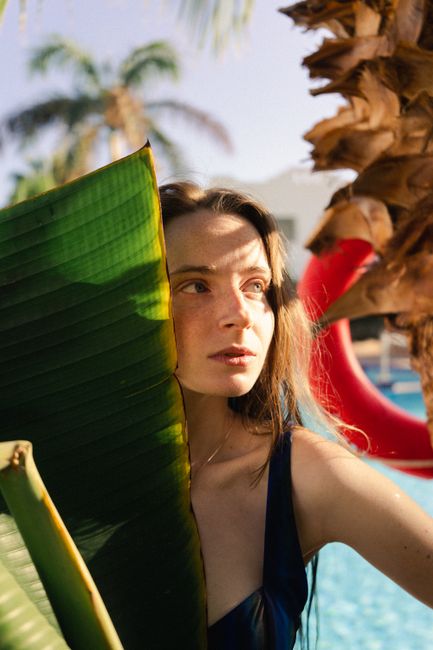 A young woman stands by a tropical pool, bathed in warm sunlight, with banana leaves nearby.