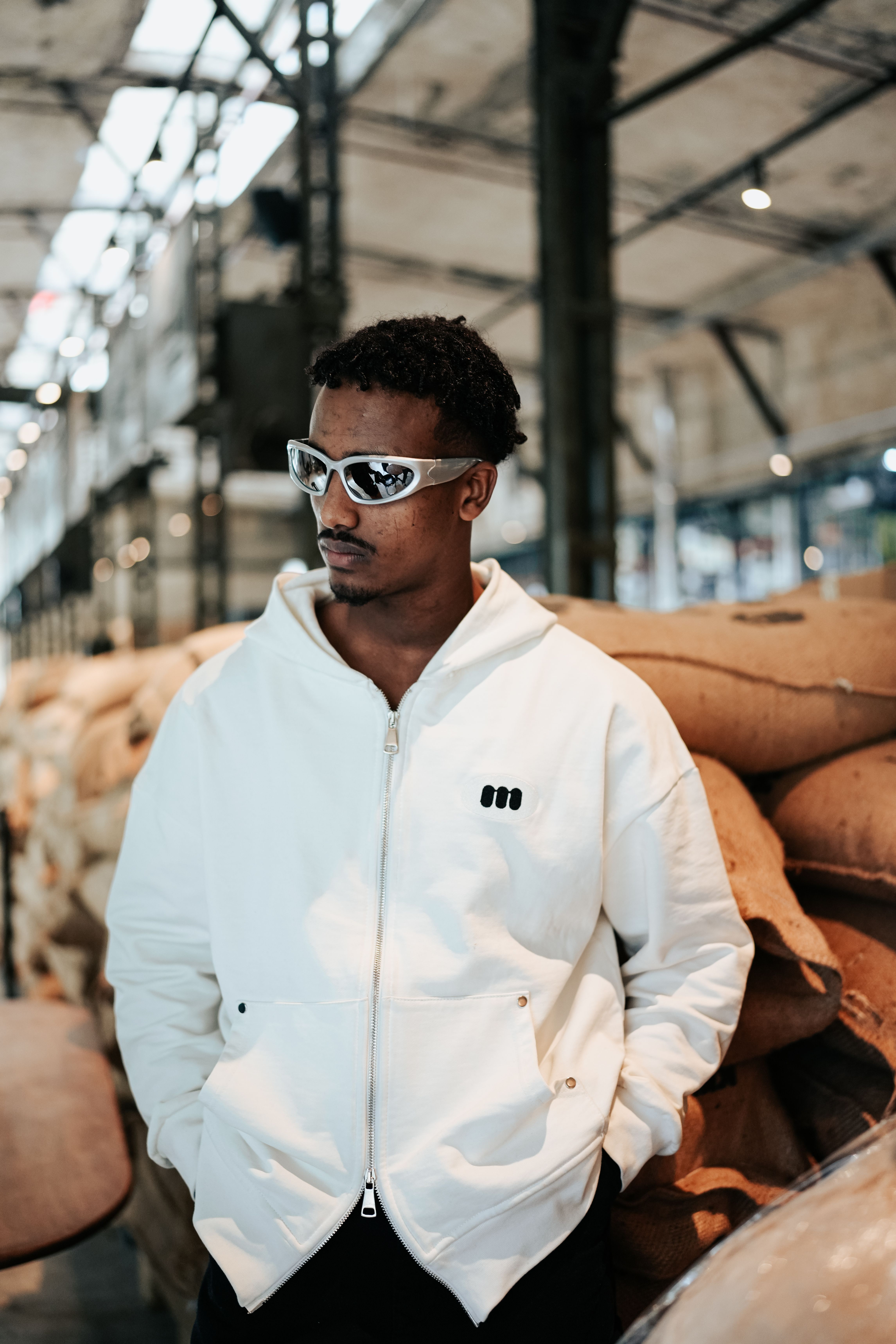 Confident young man in sunglasses and white hoodie posing in an industrial setting.