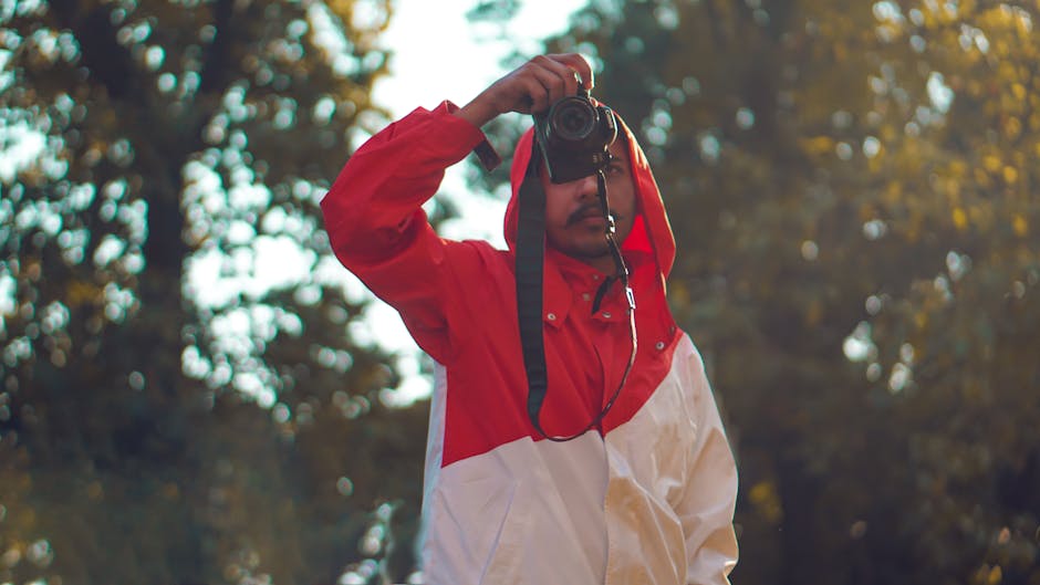 A man in a red and white jacket takes a photo outdoors surrounded by trees.