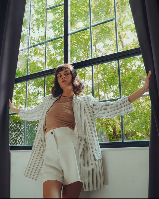 Chic woman posing by a large window with sunlight streaming in, exuding summer vibes.
