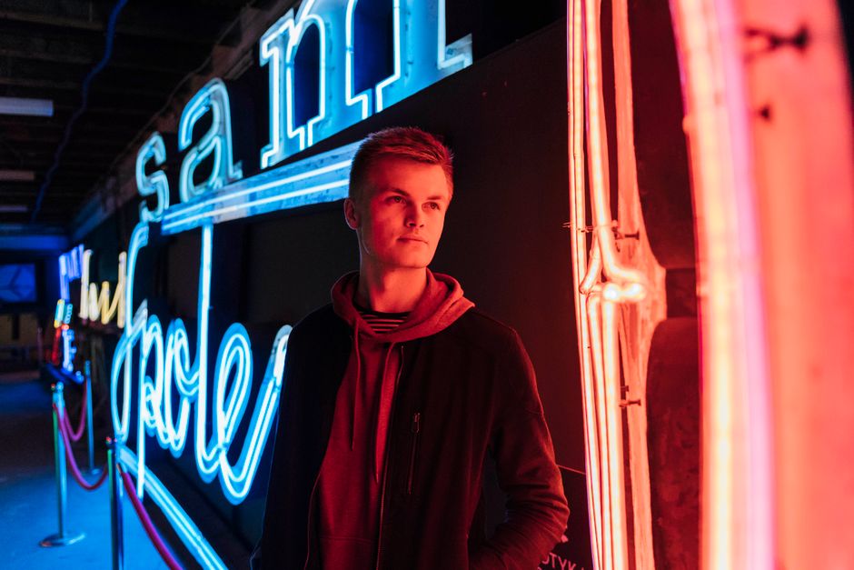 Young adult male standing beside bright neon signs, creating a vibrant contrast.