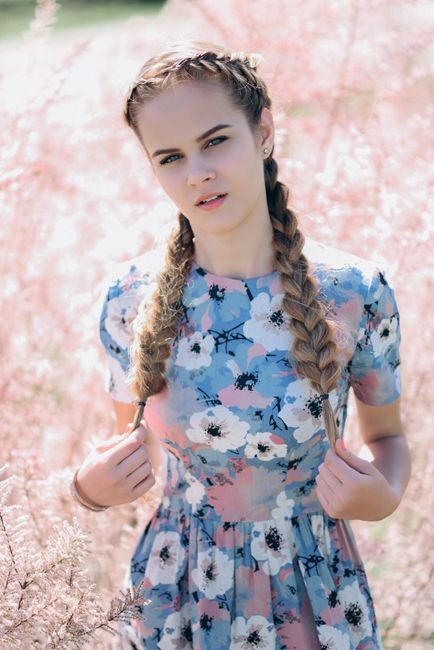 A young woman with braided hair stands in a pastel-colored meadow wearing a floral dress.
