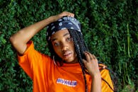 一位时尚女性，梳着编发，身着橙色上衣，站在户外郁郁葱葱的绿色背景前