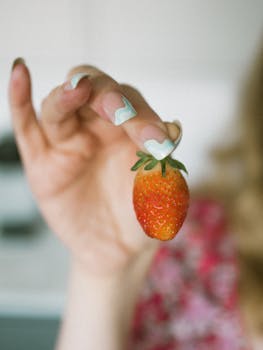 Close-up of a woman's hand holding a fresh strawberry, showcasing stylish nail art.