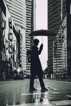 Silhouette of person with umbrella on rainy city street with skyscrapers.