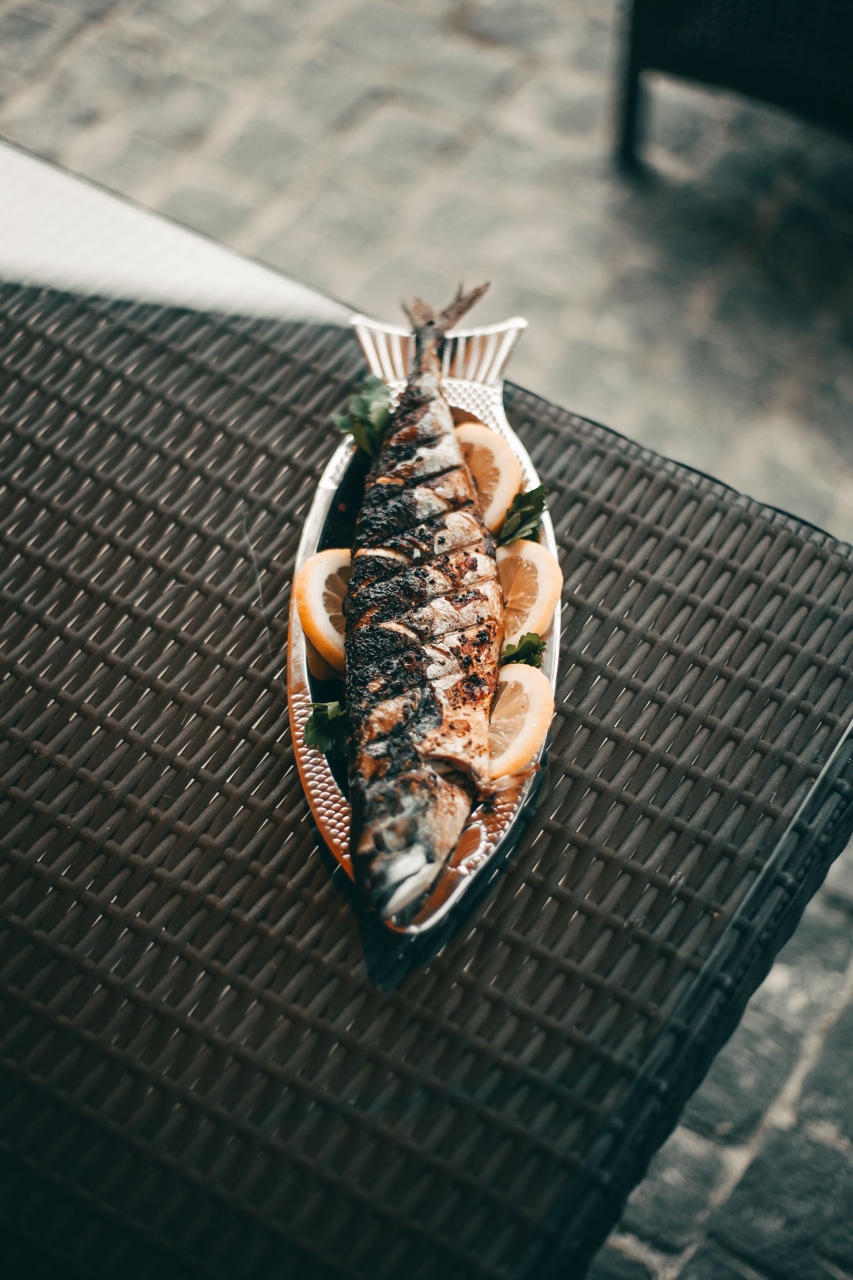 A grilled fish on a platter with lemon slices, captured in natural light on a wicker table.