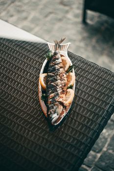 A grilled fish on a platter with lemon slices, captured in natural light on a wicker table.