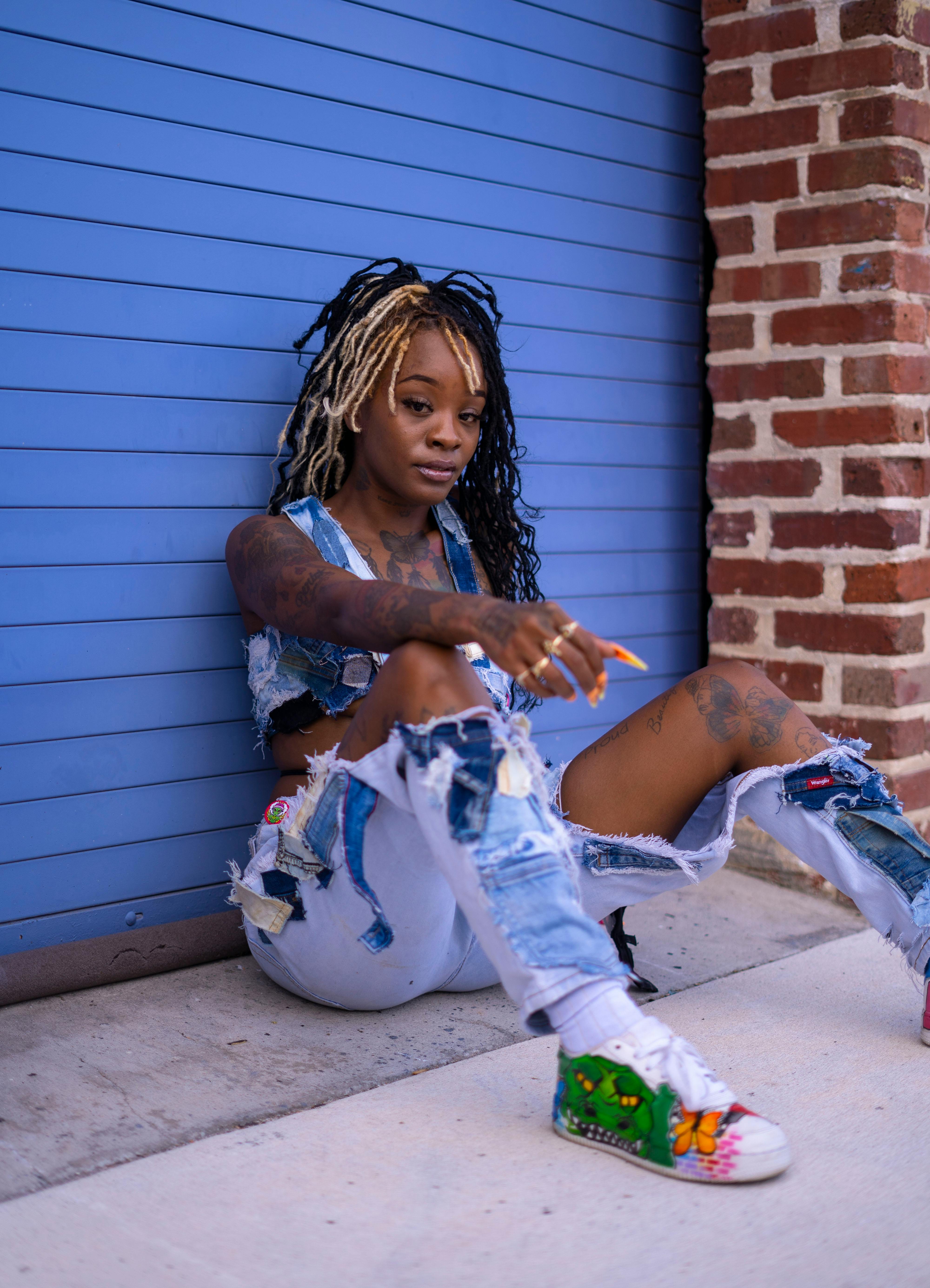 Trendy fashion portrait of a woman with dreadlocks in distressed jeans sitting against a blue door.