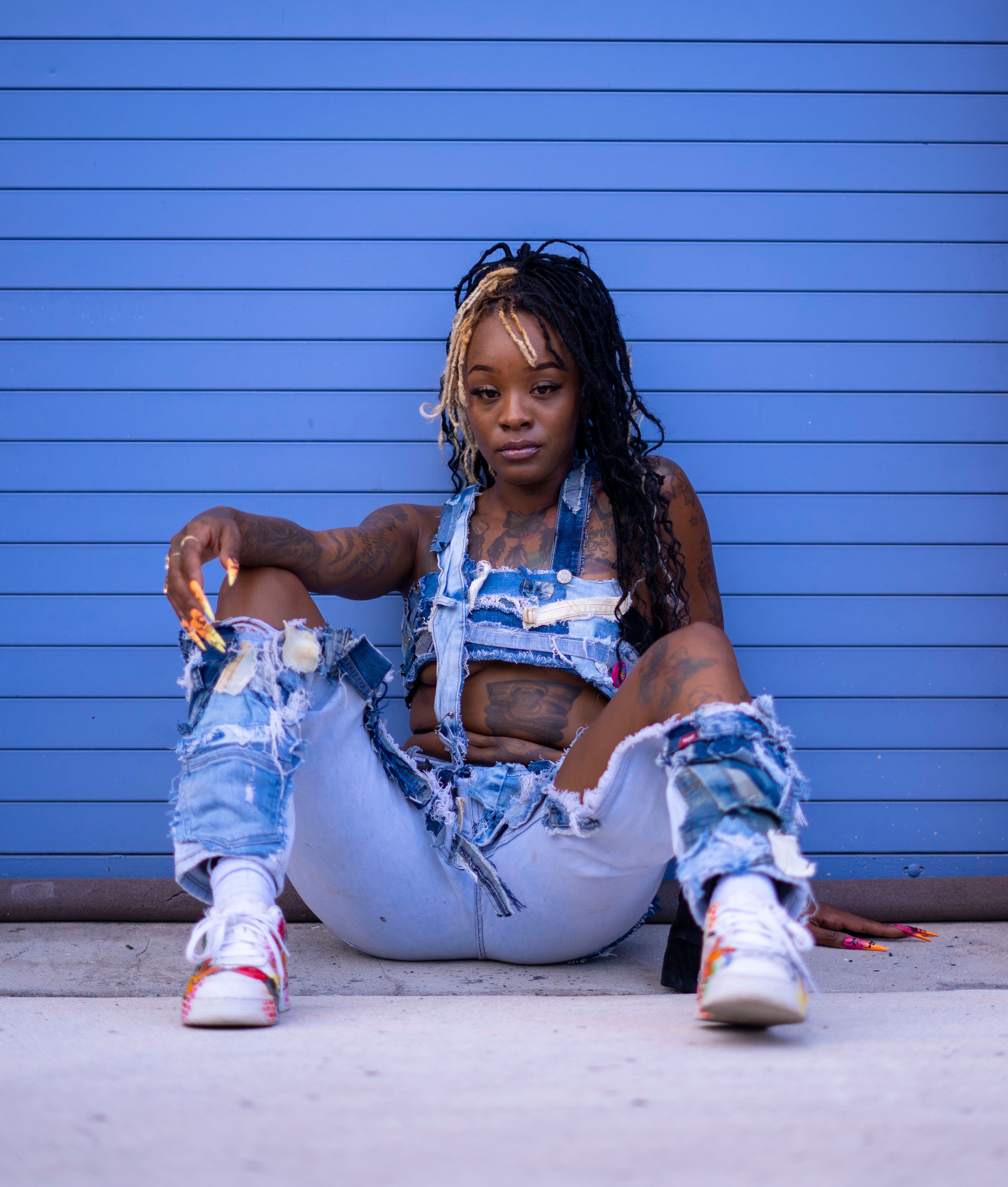 Woman sitting against a blue wall in modern distressed denim with unique style and dreadlocks.