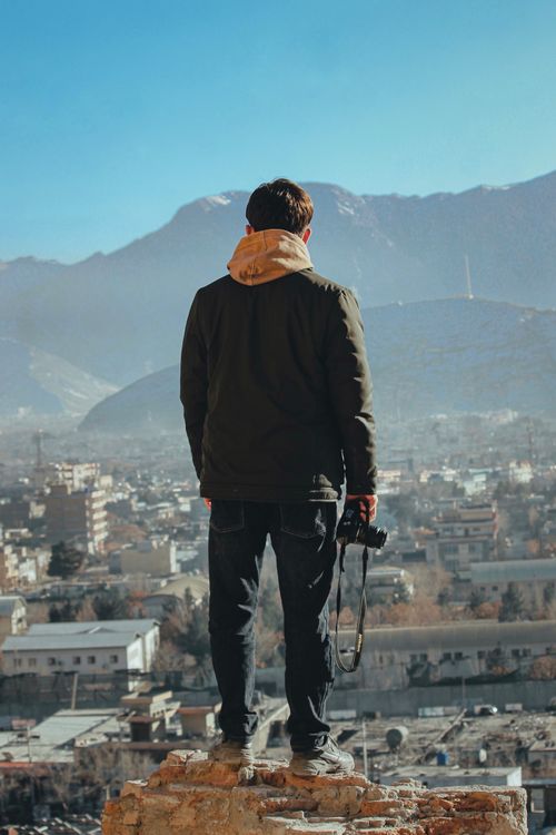 Free A man in a jacket stands atop rocks, holding a camera, overlooking Kabul cityscape. Stock Photo