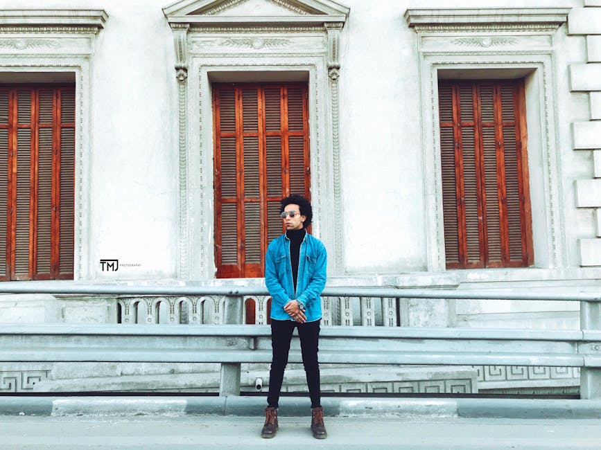 Fashionable man in denim jacket and sunglasses stands confidently against historic architecture.