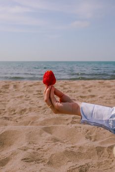 Hand holding a strawberry over sandy beach with sea in background under clear sky.