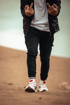 A young male showing casual style with sneakers and jacket on a sandy beach.