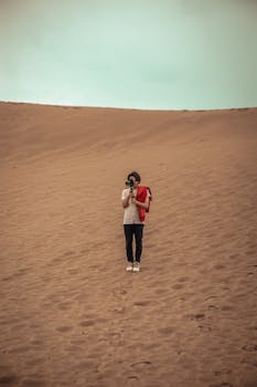 A lone man stands in a vast desert holding a camera, capturing the scenic barren landscape.