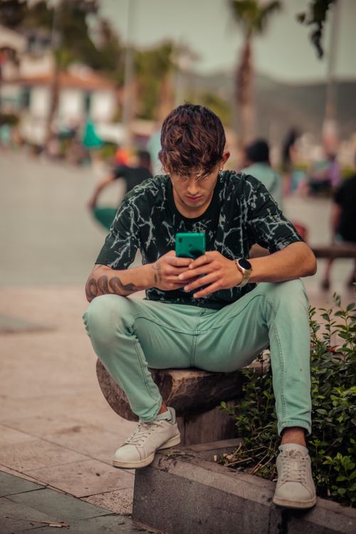 Free Young man sitting on bench outdoors, focused on smartphone. Urban lifestyle scene. Stock Photo