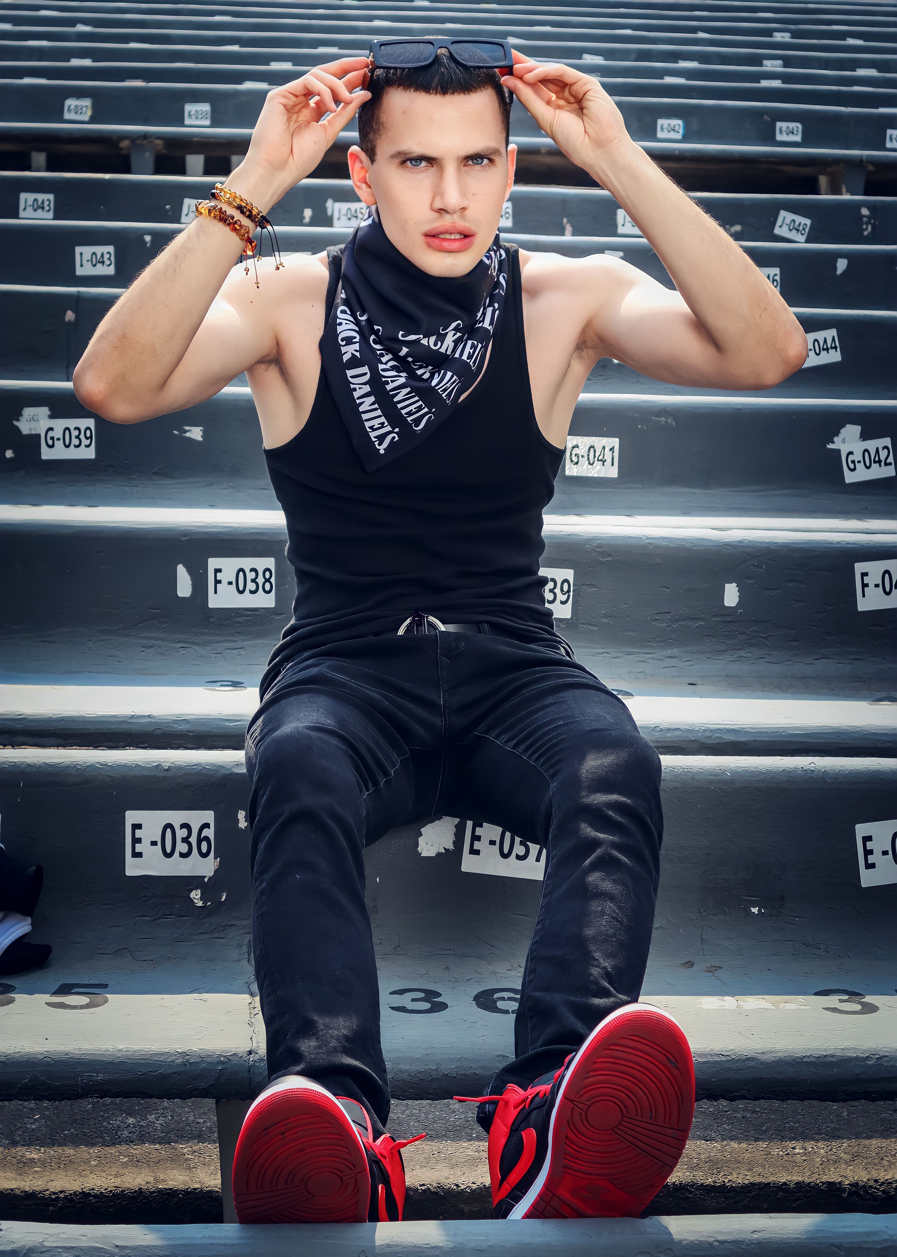 Young man wearing tank top and sunglasses, seated on stadium steps, exuding urban style.