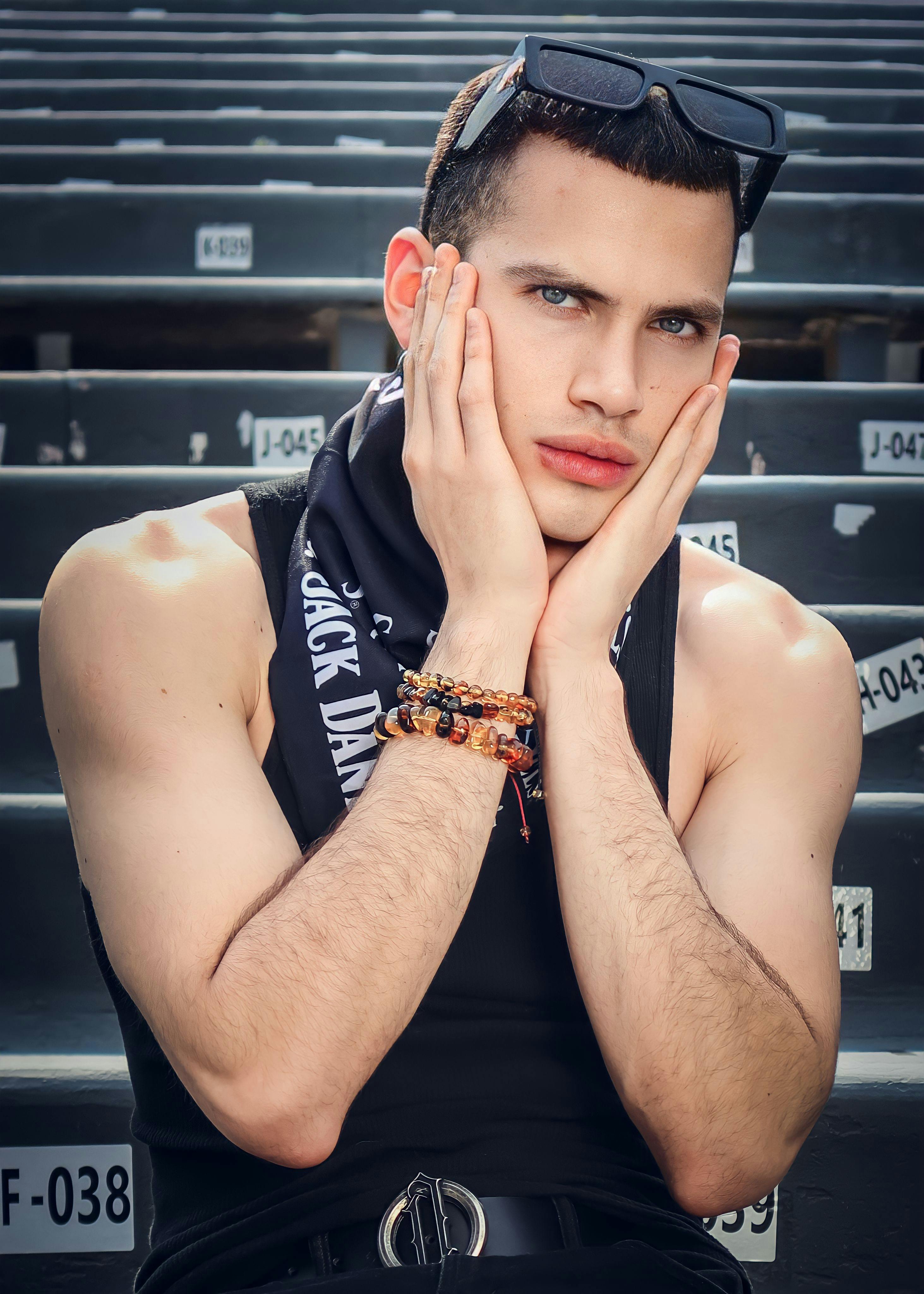 Portrait of a fashionable young man posing with hands on cheeks, wearing sunglasses and a tank top outdoors.