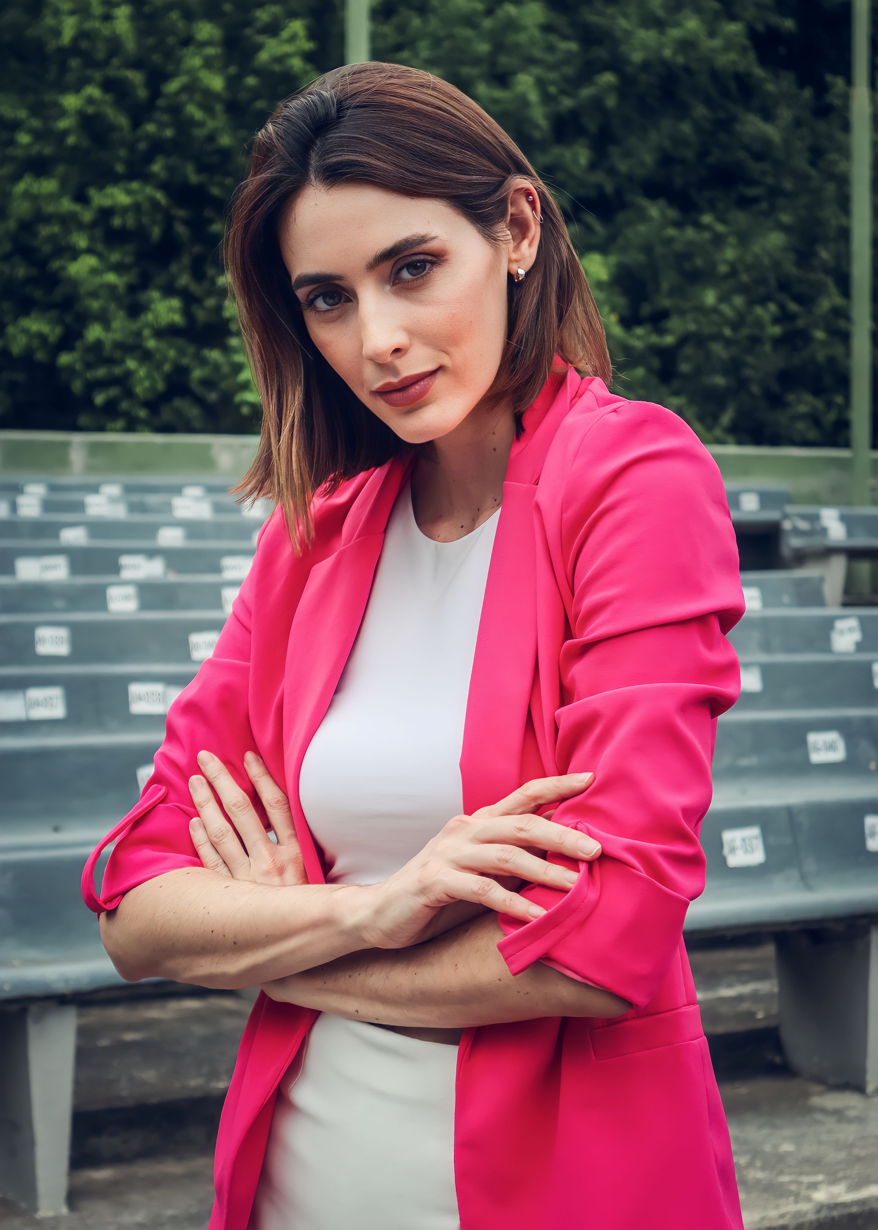 Confident woman with brown hair in a pink blazer posing with arms crossed outdoors.
