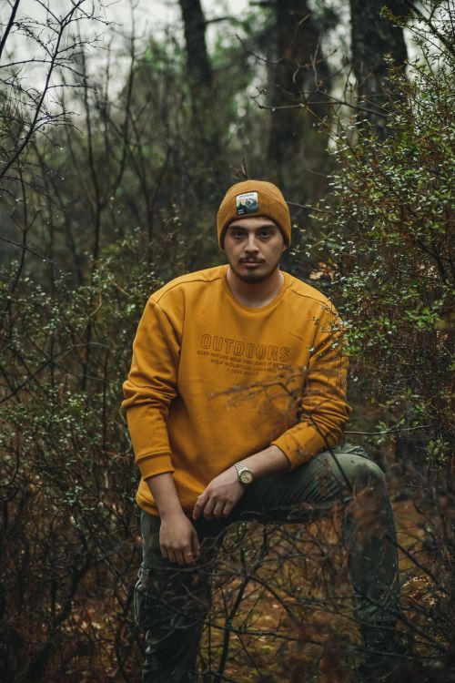 Free Portrait of a young man in a forest, wearing a yellow sweatshirt and hat, surrounded by trees. Stock Photo