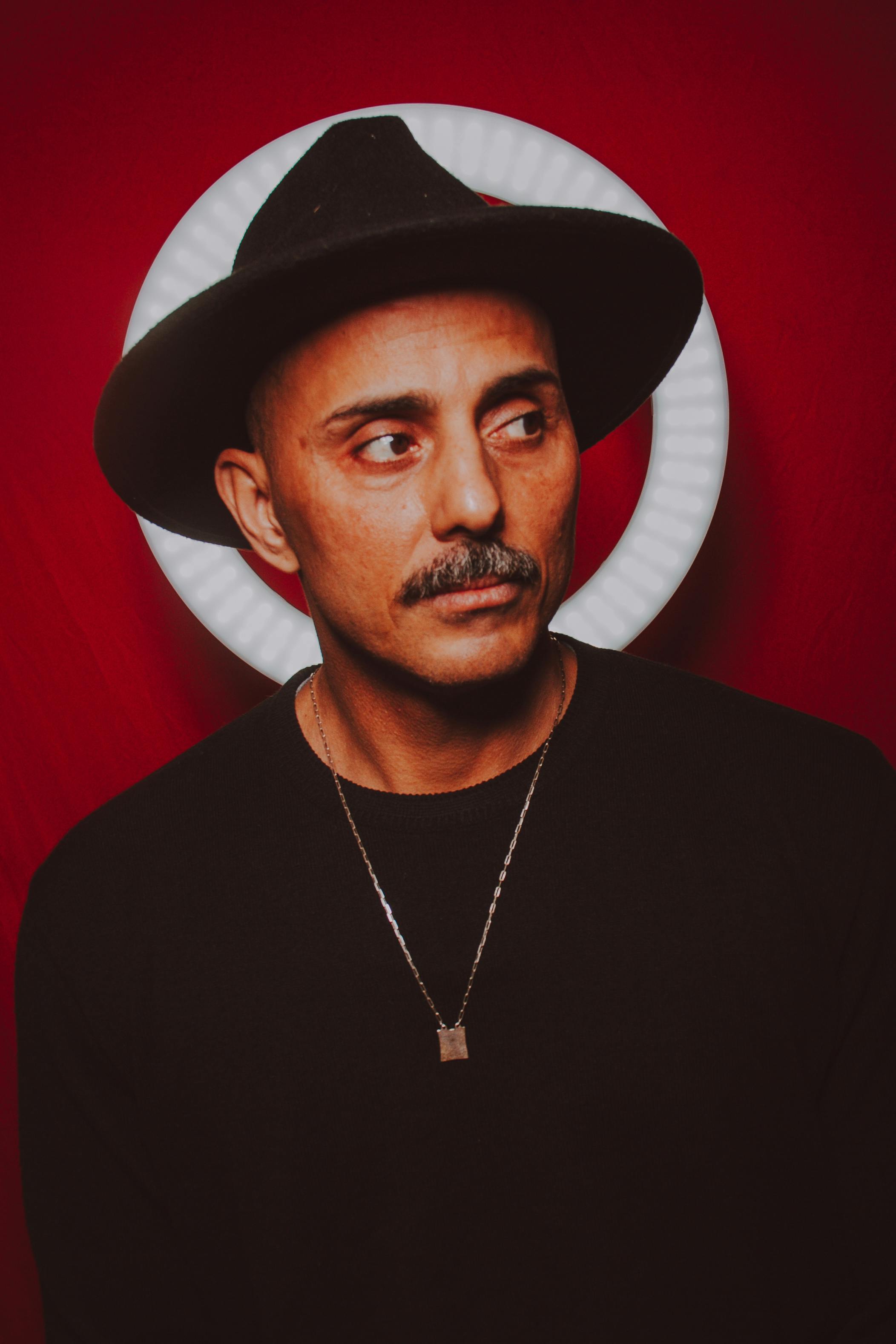 A striking studio portrait of a man wearing a hat and sweater, set against a vivid red background.