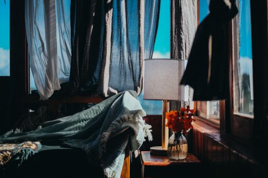 A cozy bedroom scene featuring sunlight streaming through curtains, a blanket, and a vase of flowers.