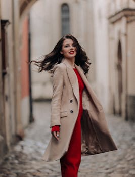 Stylish woman in a red dress and coat posing confidently on an old town street, exuding elegance.