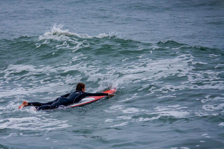 A surfer in a wetsuit paddles on a red surfboard amidst ocean waves.