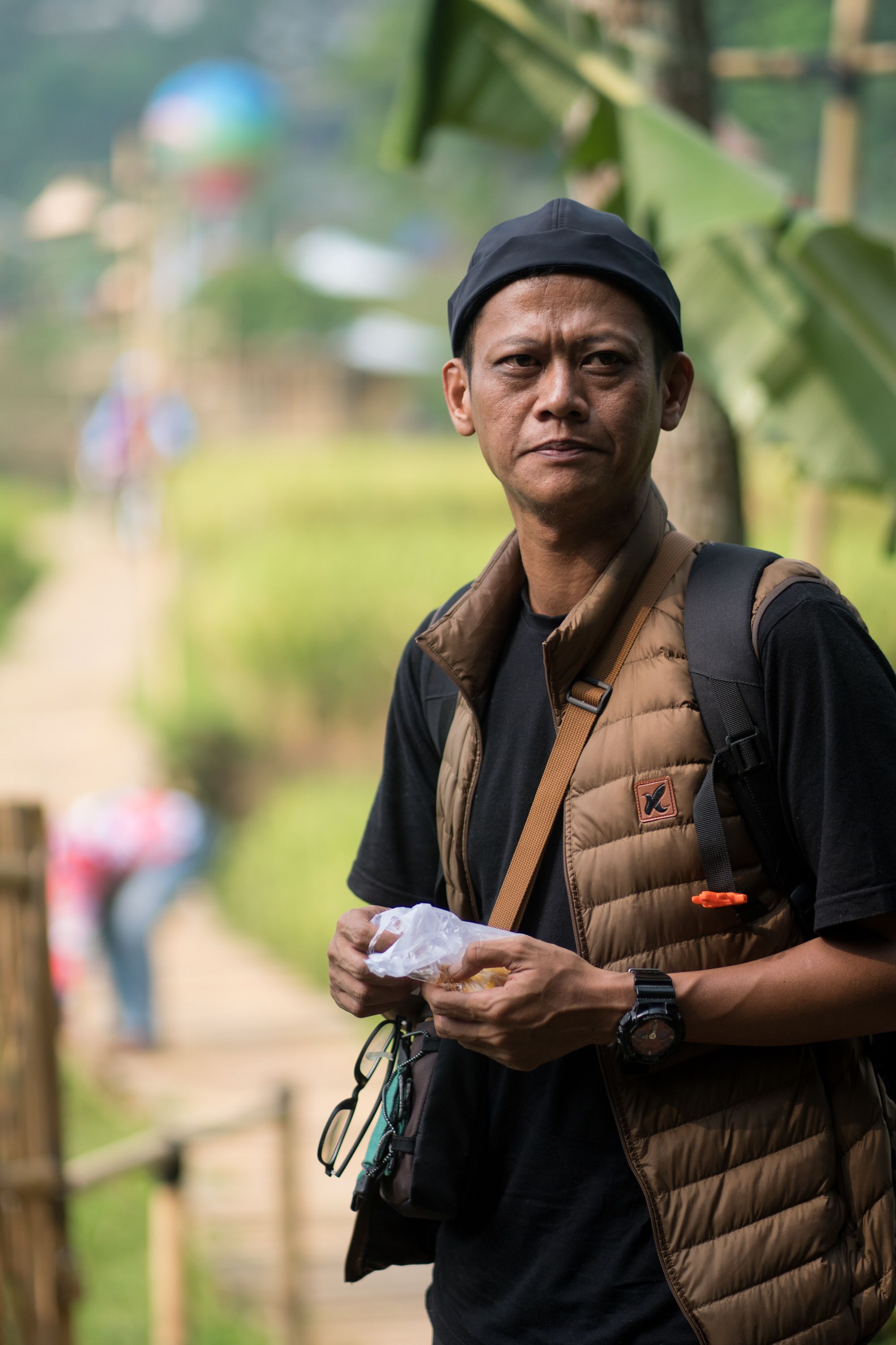Man in a vest and cap holding eyeglasses and standing outdoors on a hiking trail.